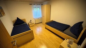 Ein Zimmer mit zwei Holzbett, einem kleinen Nachttisch mit Lampe, einem Heizkörper und einem Fenster mit Vorhängen.