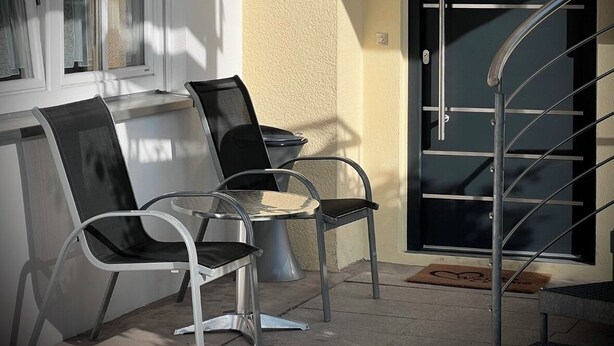 Ein moderner Außenbereich mit Metallstühlen, einem Glastisch, einer schwarzen Tür mit Metallgriff und einer Metalltreppe.