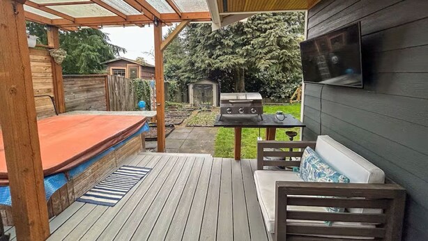 A covered outdoor patio with a wooden deck, a grill, a TV mounted on the wall, and a seating area with a cushion. - Perfect Basecamp to explore the PNW and FIFA World Cup, easy walk to lightrail (Shoreline)