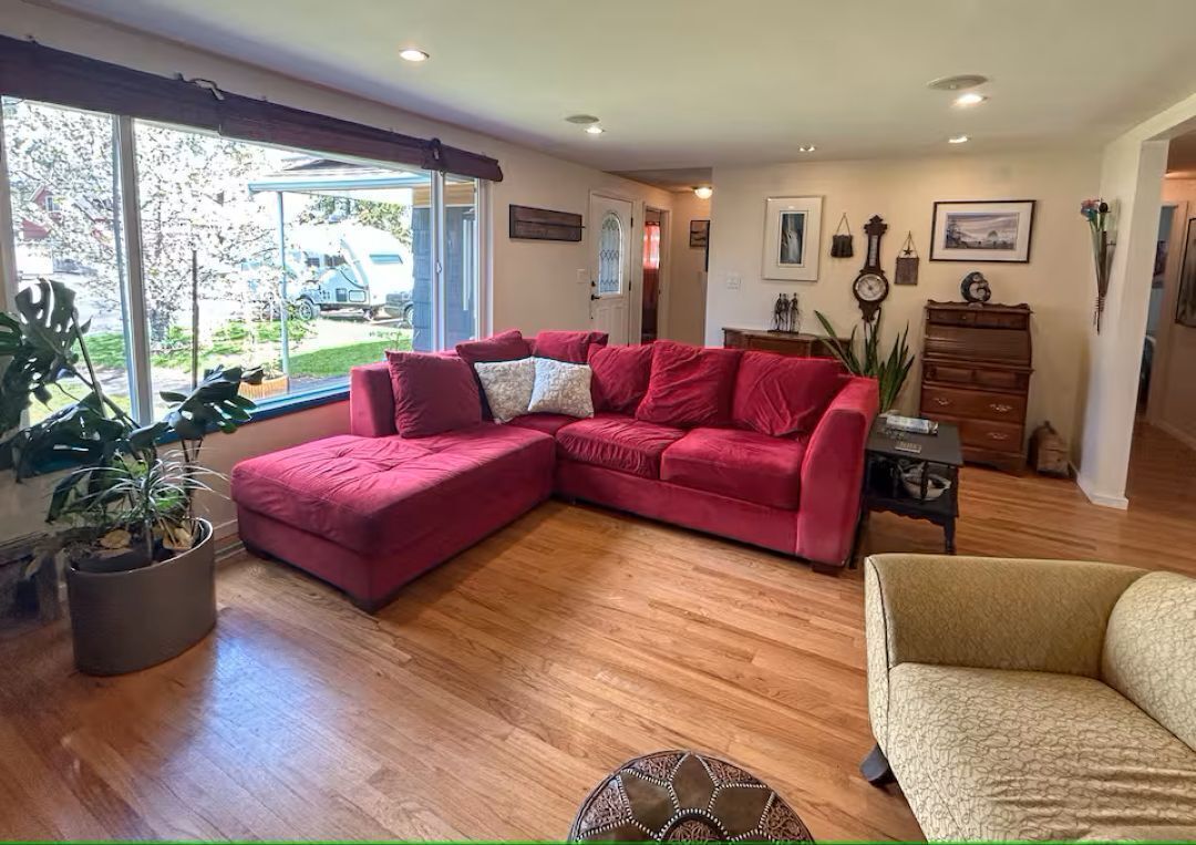 Ein Wohnzimmer mit einem roten Ecksofa, einem beigefarbenen Sessel, Holzböden, einer Topfpflanze, einer Uhr und einem Fenster mit Blick ins Freie.