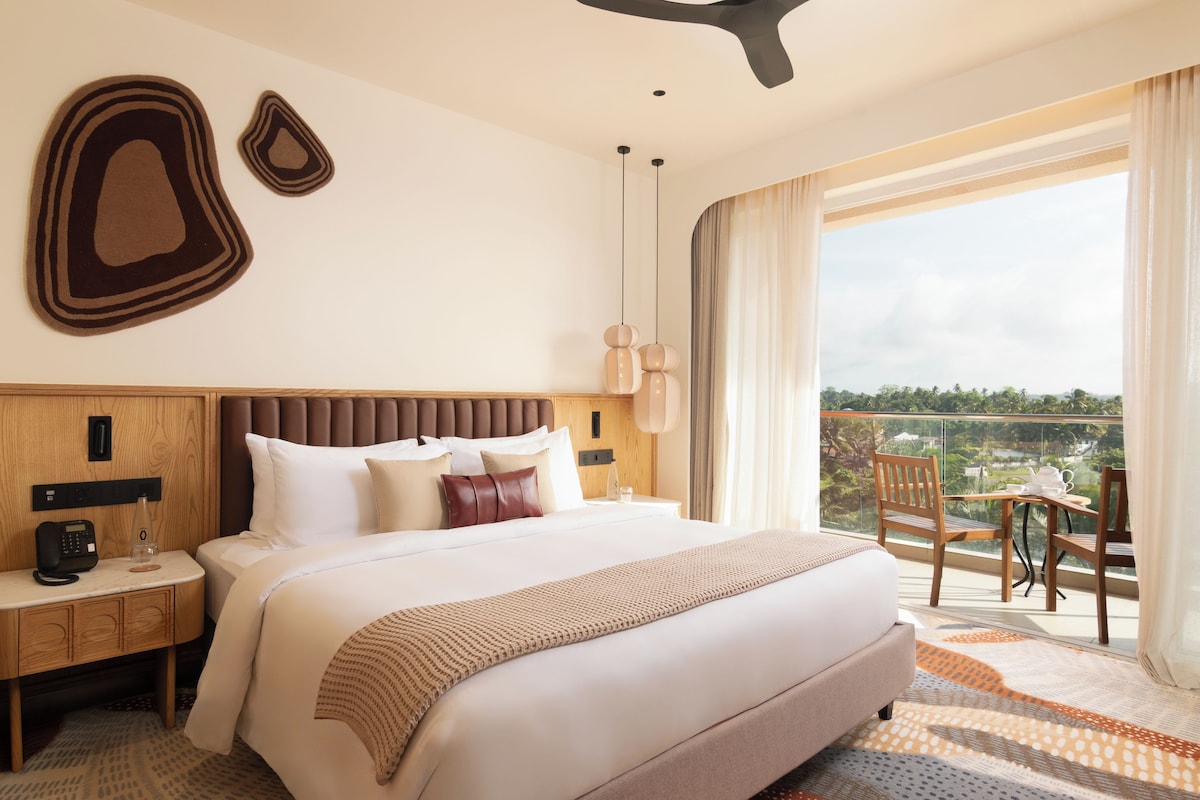 Habitación de hotel con una cama grande, una mesita de noche con un teléfono y una botella, una ventana con vistas a zonas verdes y un balcón con mesa y sillas.