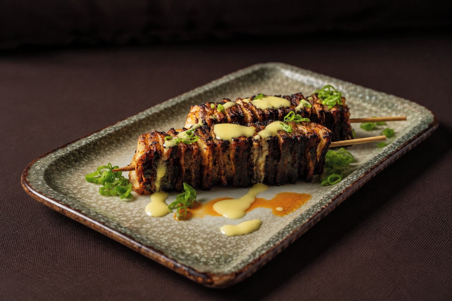Un plato con brochetas de carne a la parrilla, salsa cremosa y guarnición verde sobre un plato texturizado.