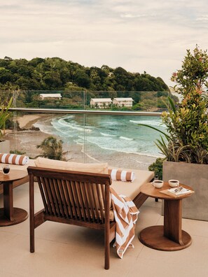 Un patio frente a la playa con una silla y mesa de madera, vista al océano y una casa al fondo.