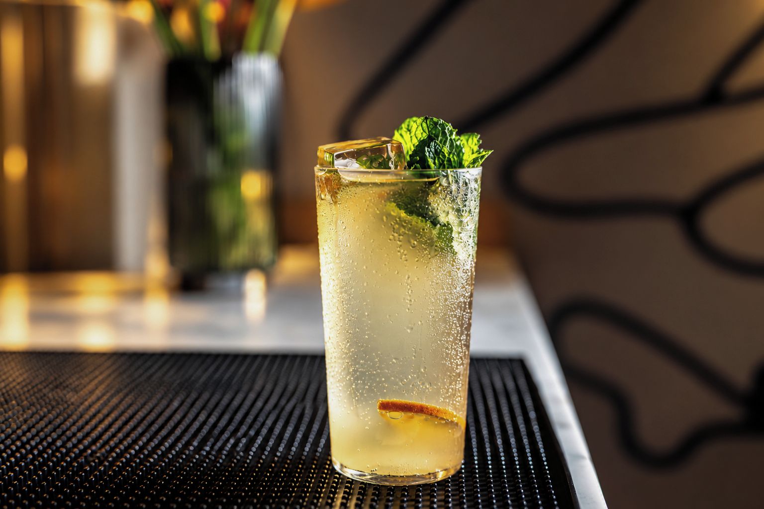 A tall glass with a beverage, garnished with a mint leaf, on a table with a black textured surface.