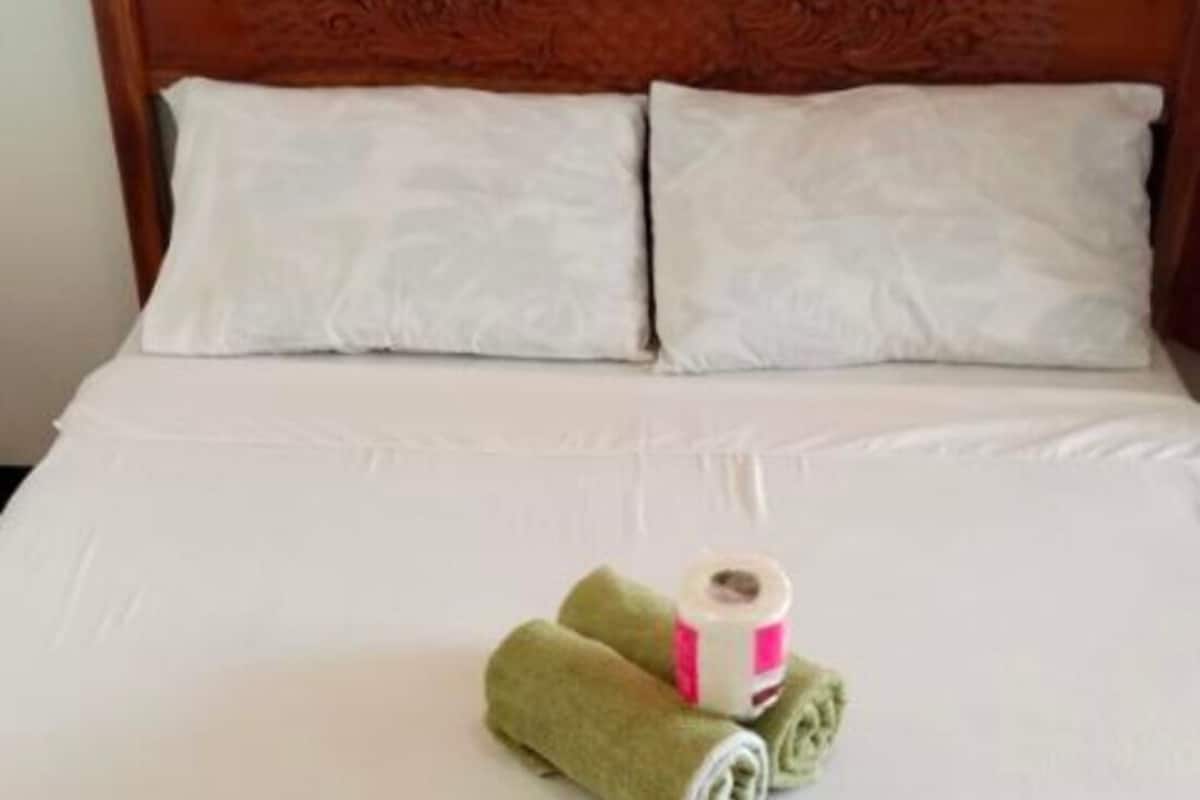 Un lit avec du linge de lit blanc, deux oreillers et une serviette verte enroulée, accompagnée d’une bobine de fil.