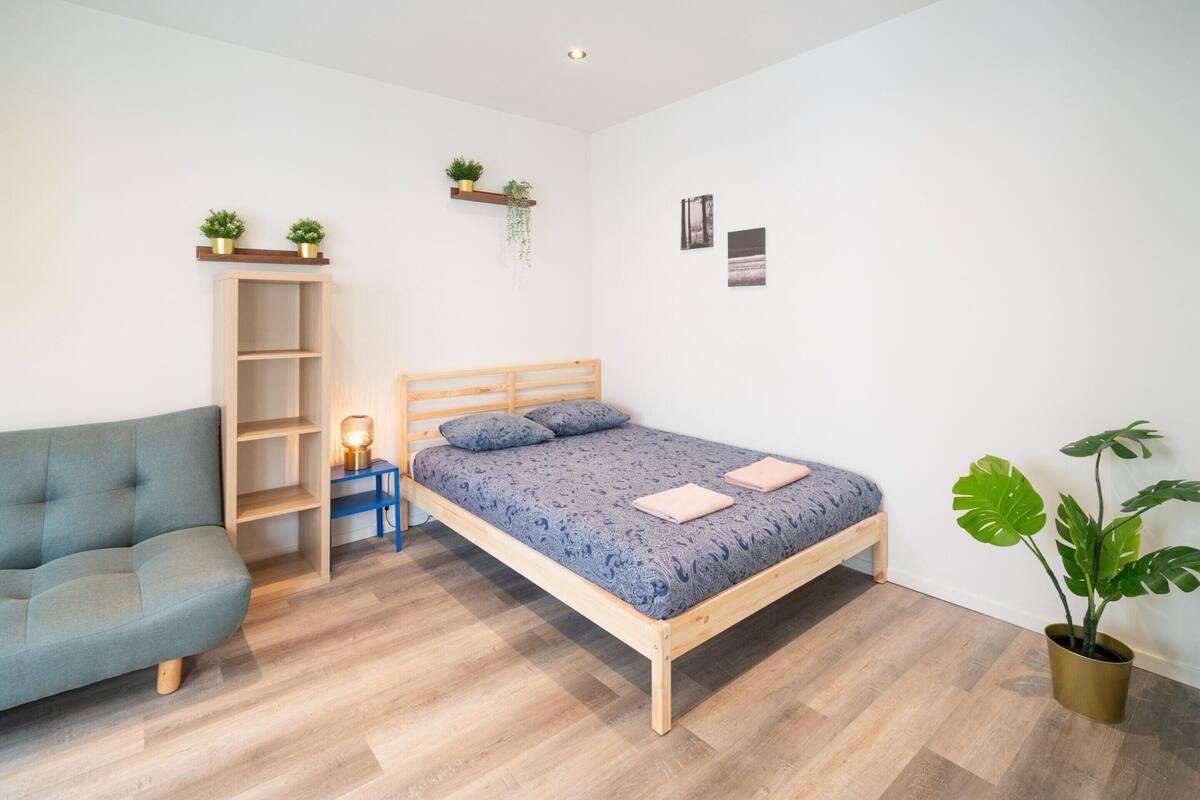 A bedroom with a bed, a sofa, a bedside table, a lamp, a shelf with plants, and a potted plant.