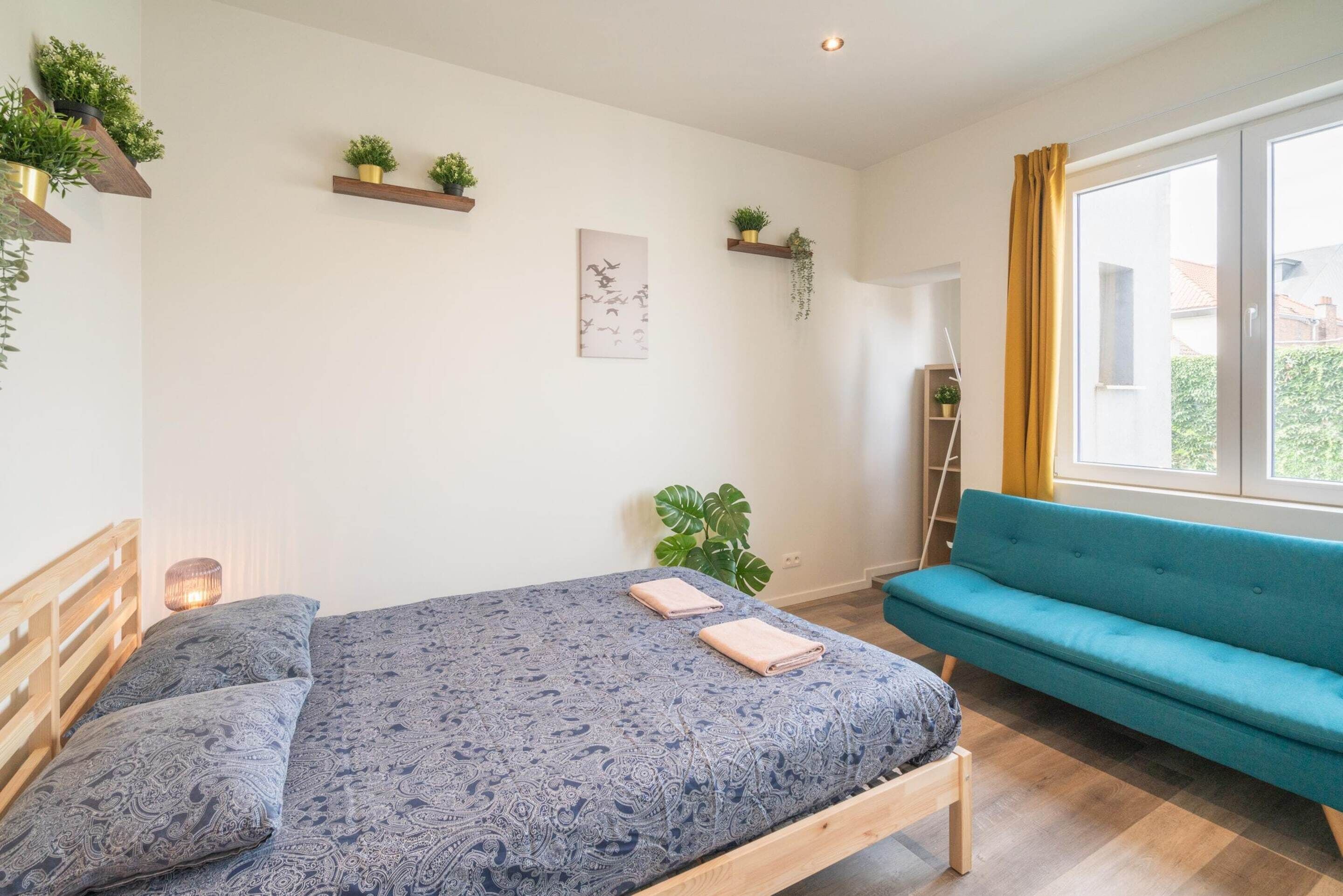 Une chambre à coucher avec un lit, un canapé bleu, une fenêtre avec des rideaux et des étagères murales ornées de plantes.