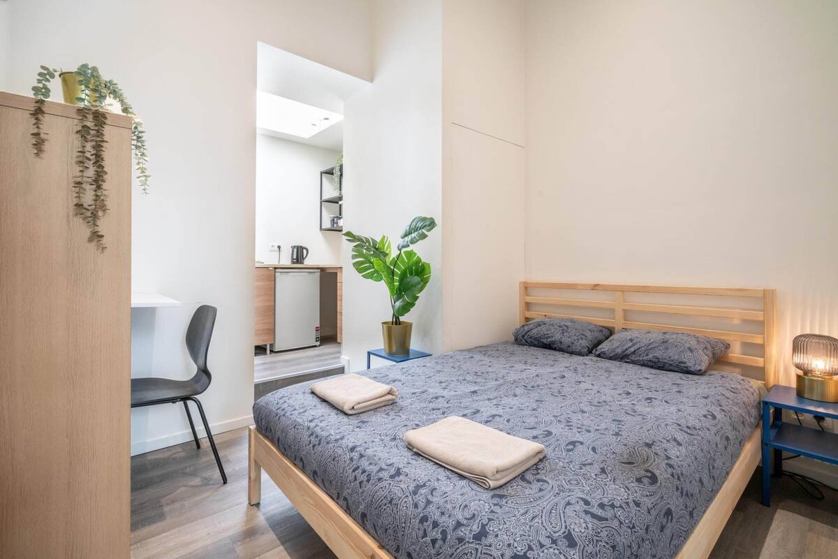 A bedroom with a bed, bedside table, chair, desk, and potted plant.