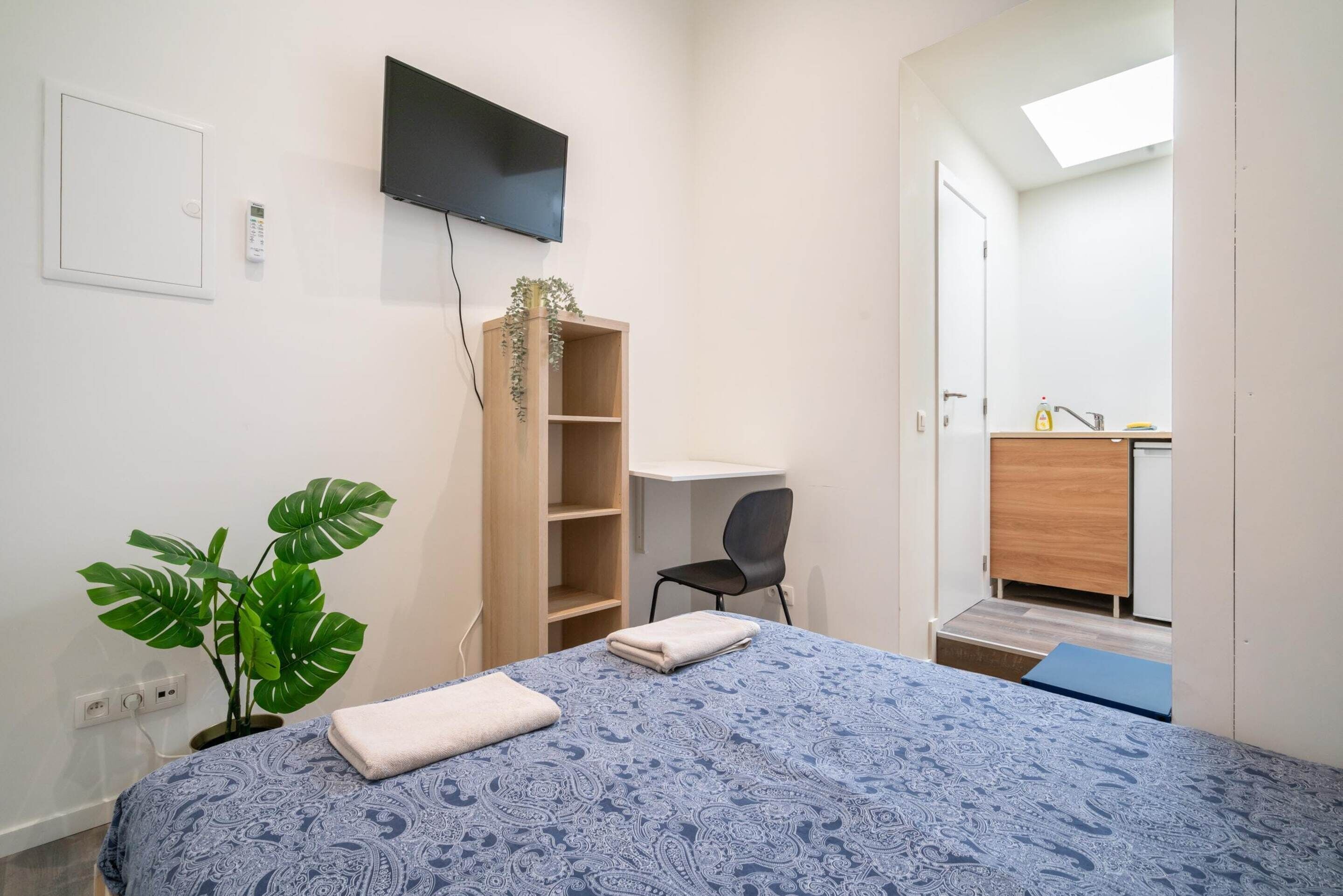 Un dormitorio con cama, escritorio, silla, televisor, planta y un baño visible a través de una puerta abierta.
