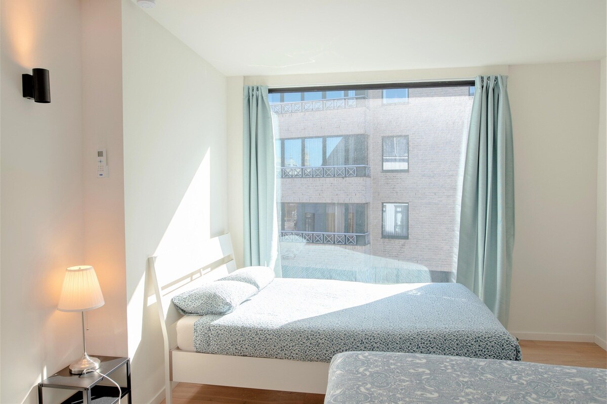 A bedroom with a bed, a bedside table with a lamp, a window with curtains, and a view of a building outside.