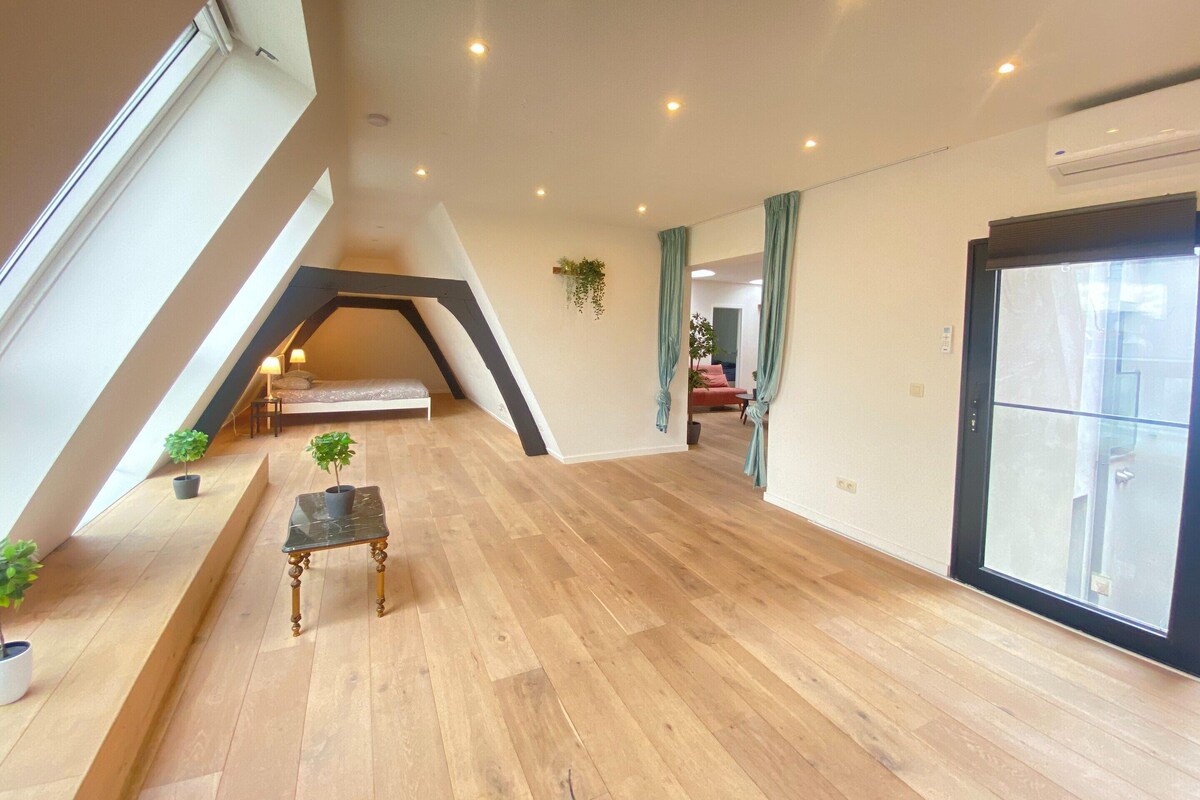 A spacious loft bedroom with a sloped ceiling, a bed, a small table, and a potted plant.