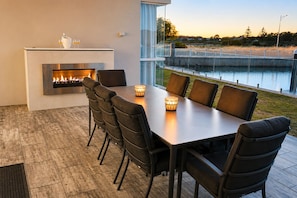 A modern outdoor dining area with a built-in fireplace, a glass railing overlooking a body of water, and a view of the landscape. - Winter Mornings on the Canal - A Waterside Paradise (Geographe)