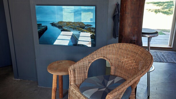 Une chaise en osier avec un coussin, un petit tabouret et un tableau encadré représentant un paysage côtier.