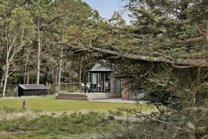 Una casa moderna con puerta de cristal, una terraza con sillas y una caseta pequeña en un área boscosa.