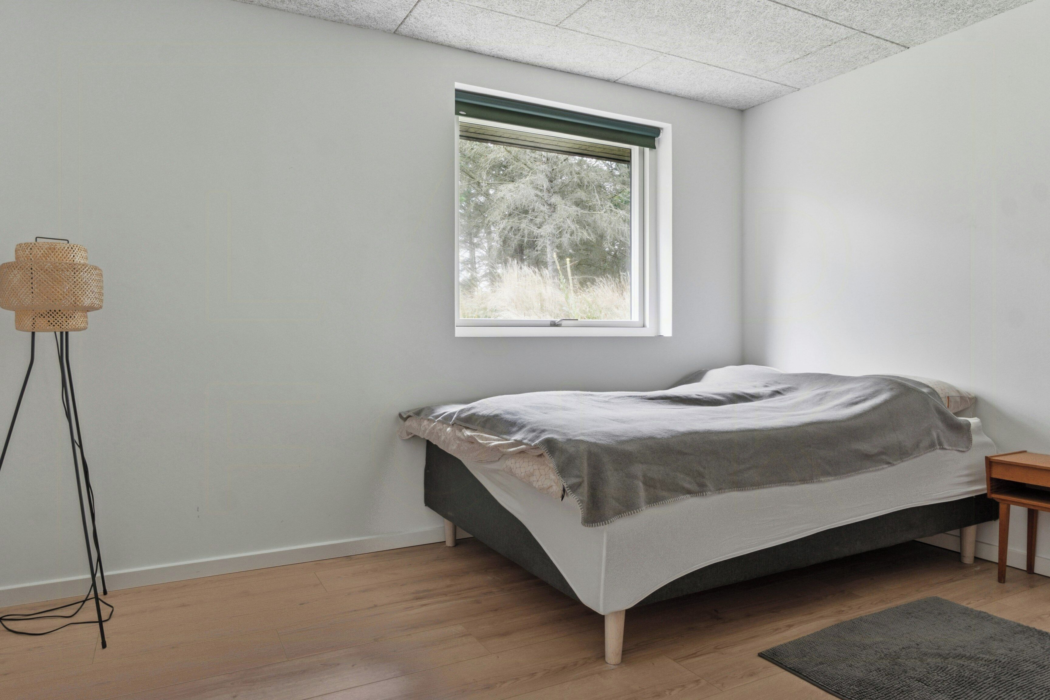 A bedroom with a bed, a window, a floor lamp, and a small wooden nightstand.