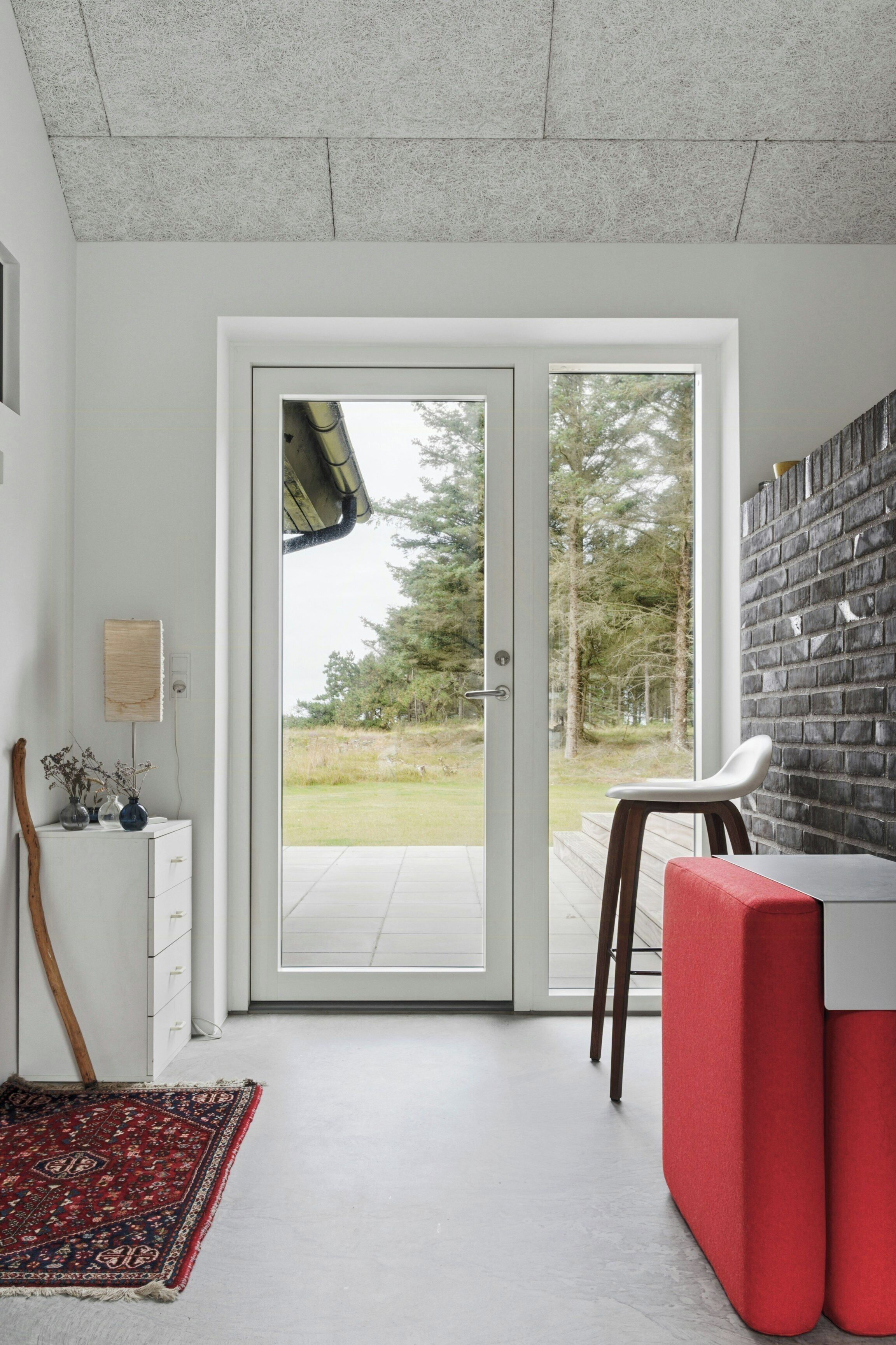 Een modern interieur met een witte muur, een rode bank, een wit bureau en uitzicht door een glazen deur op een buitenruimte.