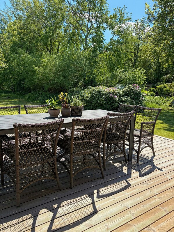 Een buitenruimte met een houten terras, rieten stoelen en een tafel.