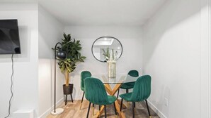 A dining area with a glass table, four green chairs, a potted plant, a round mirror, and a TV mounted on the wall. - Orchards Delight | Near Airport | Sleeps 4 | WiFi (Edmonton)