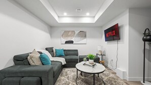 A modern living room with a grey sofa, a round coffee table, and a flat-screen TV mounted on the wall. - Orchards Delight | Near Airport | Sleeps 4 | WiFi (Edmonton)