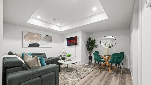 A modern living room with a grey sofa, a coffee table, a TV mounted on the wall, and a dining area with a round table and green chairs. - Orchards Delight | Near Airport | Sleeps 4 | WiFi (Edmonton)