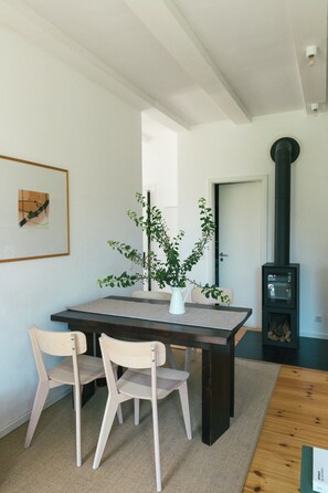 A dining area with a table, two chairs, a vase with a plant, a framed picture on the wall, and a wood-burning stove. - Tower villa apartment with pergola at Mühlenberg (Werder (Havel))