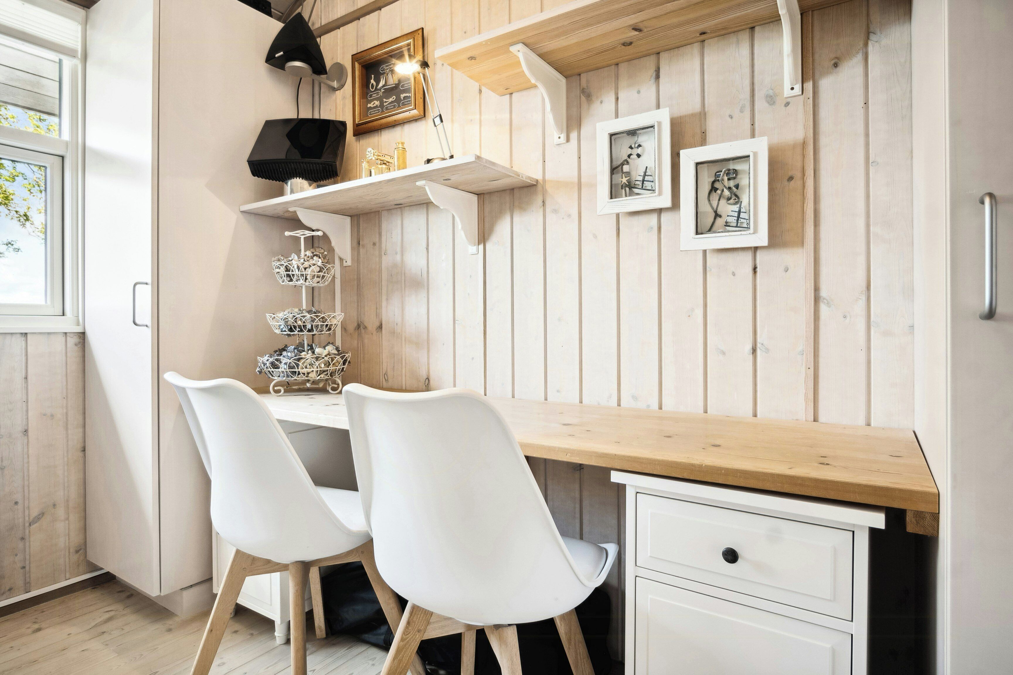 A wooden desk with two white chairs, a shelf with decorative items, and framed pictures on the wall.