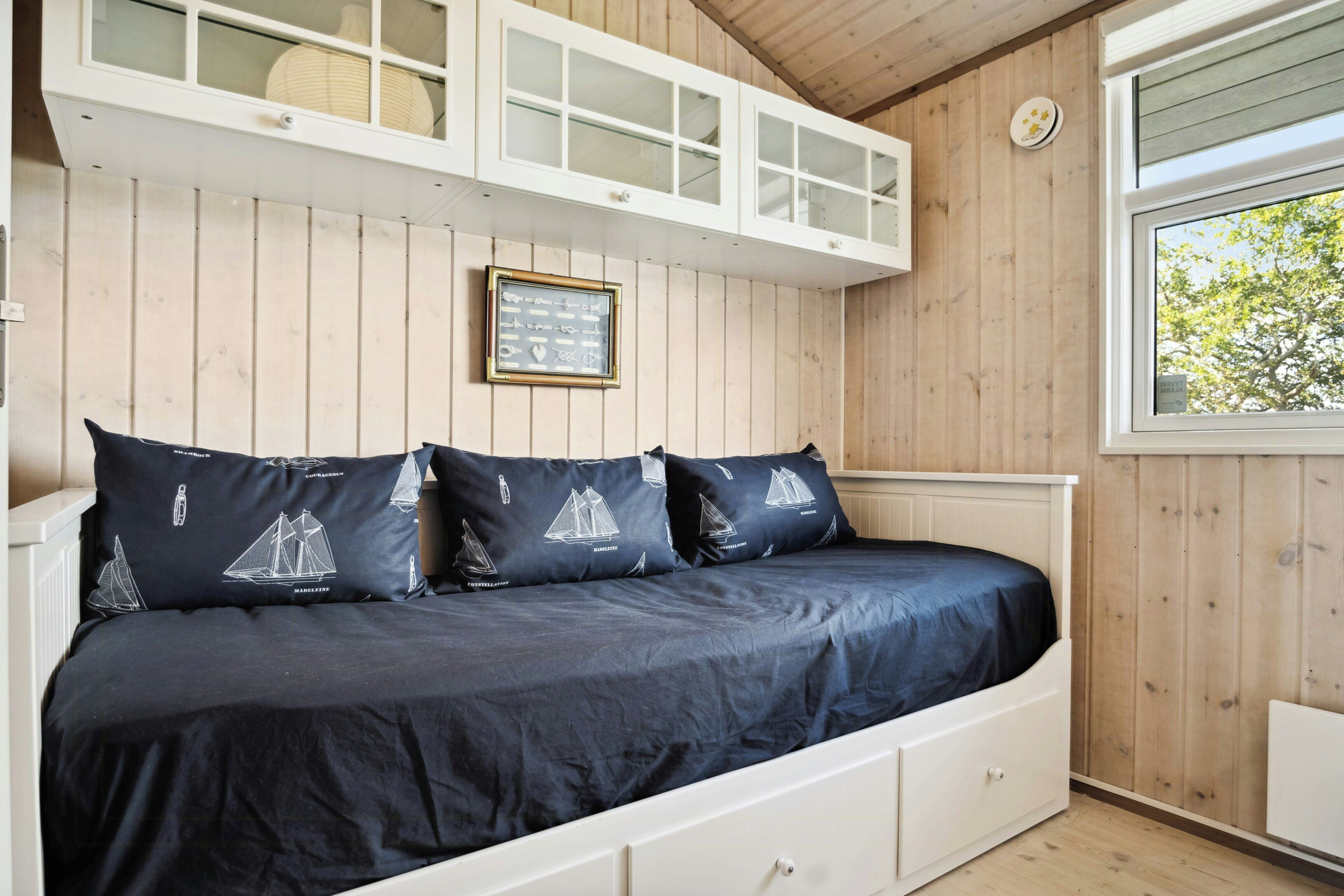 Een bed met een donkerblauwe spreidek en kussens met zeilbootmotieven, een wit hoofdeinde en een raam met uitzicht op bomen.
