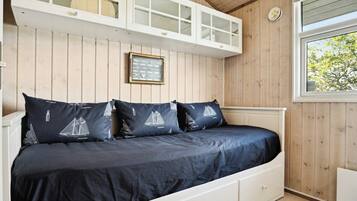 Een bed met een donkerblauwe spreidek en kussens met zeilbootmotieven, een wit hoofdeinde en een raam met uitzicht op bomen.