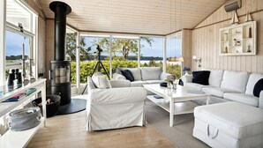 A cozy room with a fireplace, white furniture, and large windows offering a view of greenery.