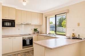 A modern kitchen with light-colored cabinets, a built-in microwave, a stove, and a window with a view of greenery. - The Sunny Brick Cottage | 3BR Bright Home w/ Patio - Holiday Home (Rochedale South)