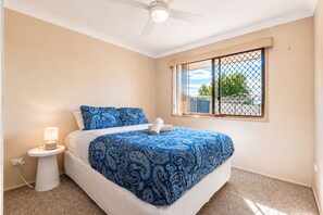 A bedroom with a bed, a nightstand, a lamp, a window with a view, and a ceiling fan. - The Sunny Brick Cottage | 3BR Bright Home w/ Patio - Holiday Home (Rochedale South)