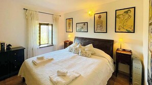 Uma cama bem arrumada com almofadas e uma toalha dobrada, uma mesinha de cabeceira com abajur, uma janela com cortinas e obras de arte emolduradas na parede.