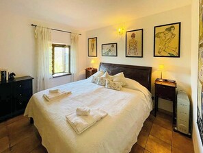 Ein ordentlich bezogenes Bett mit Kissen und einem gefalteten Handtuch, ein Nachttisch mit Lampe, ein Fenster mit Vorhängen sowie gerahmte Kunstwerke an der Wand.