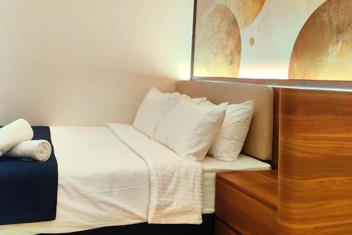 A neatly made bed with a white comforter and pillows, a wooden nightstand with a lamp, and a large abstract painting on the wall.