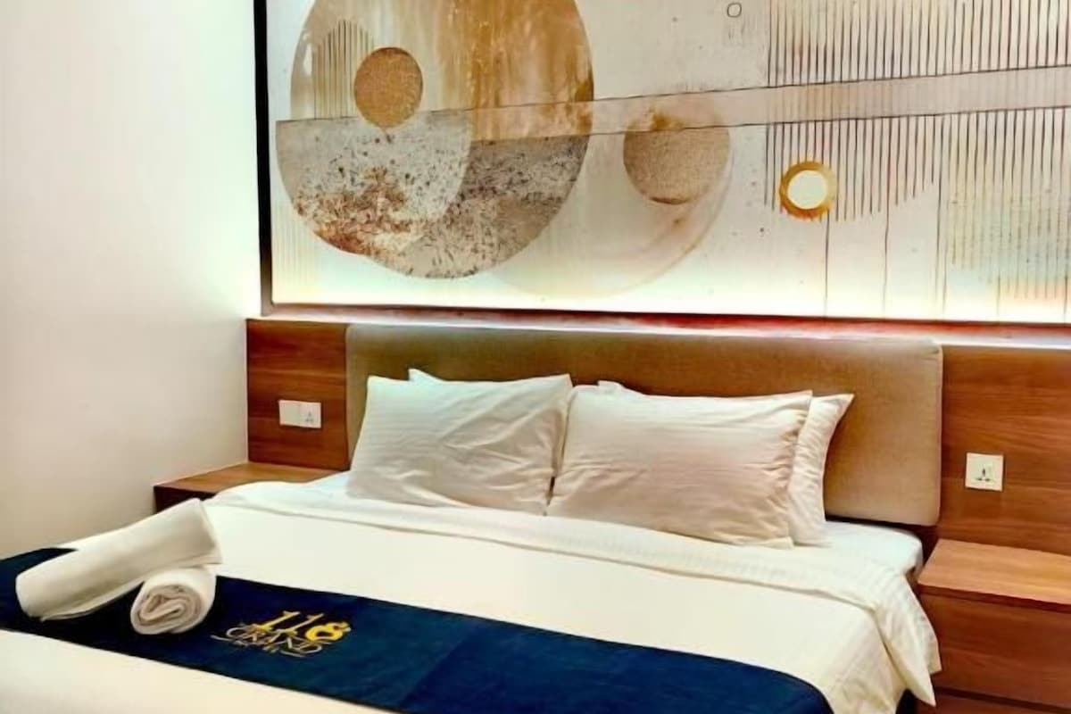 A hotel room with a large bed, a headboard with a wooden panel, a wall-mounted abstract artwork, and a neatly made bed with a blue blanket.