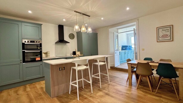 Une cuisine moderne avec un îlot central, des tabourets de bar, une table à manger avec des chaises et une horloge visible au mur.