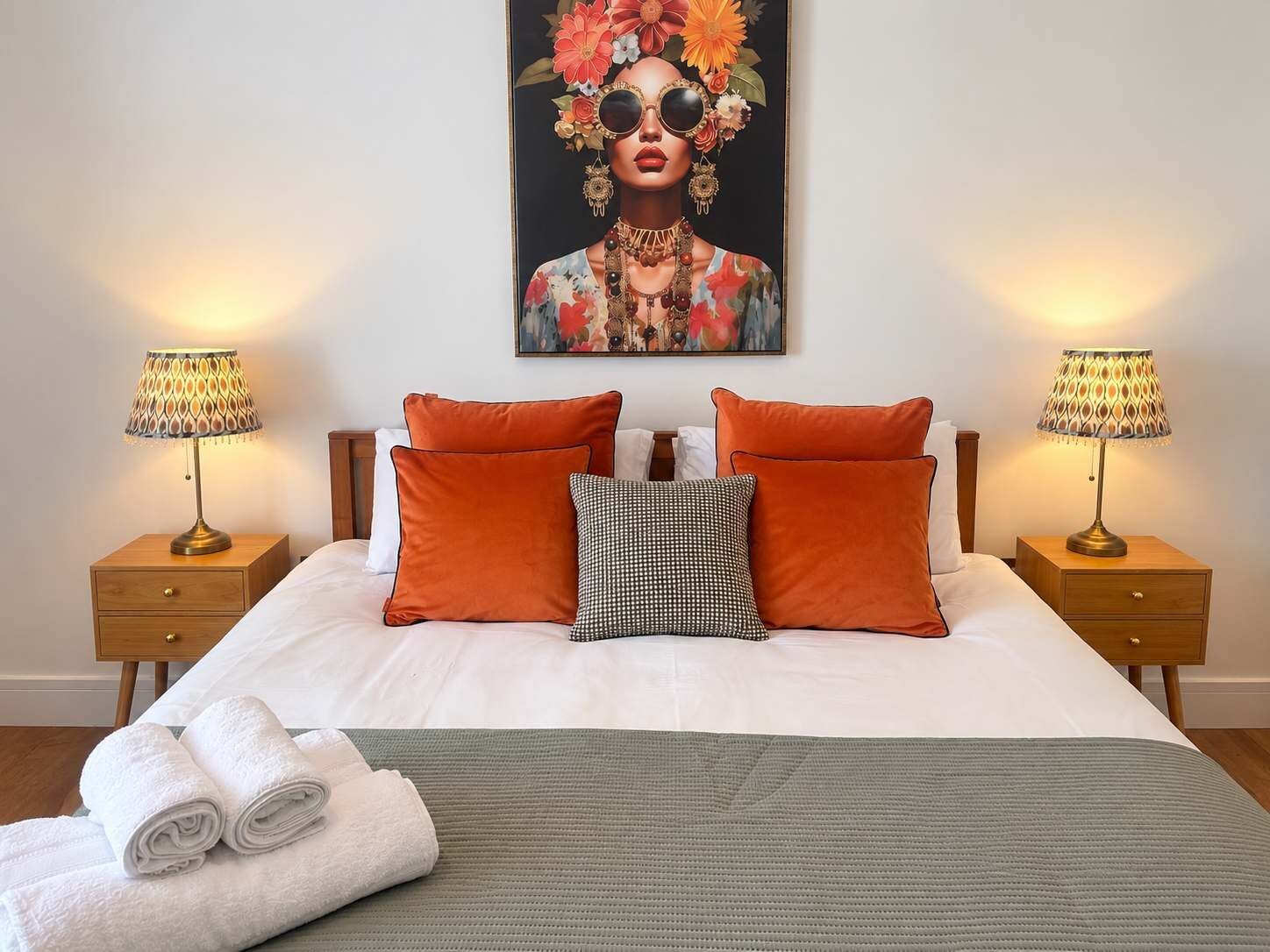 Una cama bien hecha con una colcha verde, dos almohadas naranjas, una almohada a cuadros blanco y negro, y dos lámparas de noche sobre mesitas de madera.