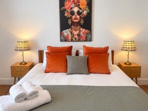 Una cama bien hecha con una colcha verde, dos almohadas naranjas, una almohada a cuadros blanco y negro, y dos lámparas de noche sobre mesitas de madera.