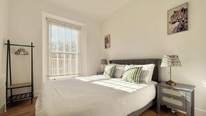 Une chambre à coucher comprenant un lit, des tables de chevet, une fenêtre avec des stores, un radiateur et un tableau accroché au mur.