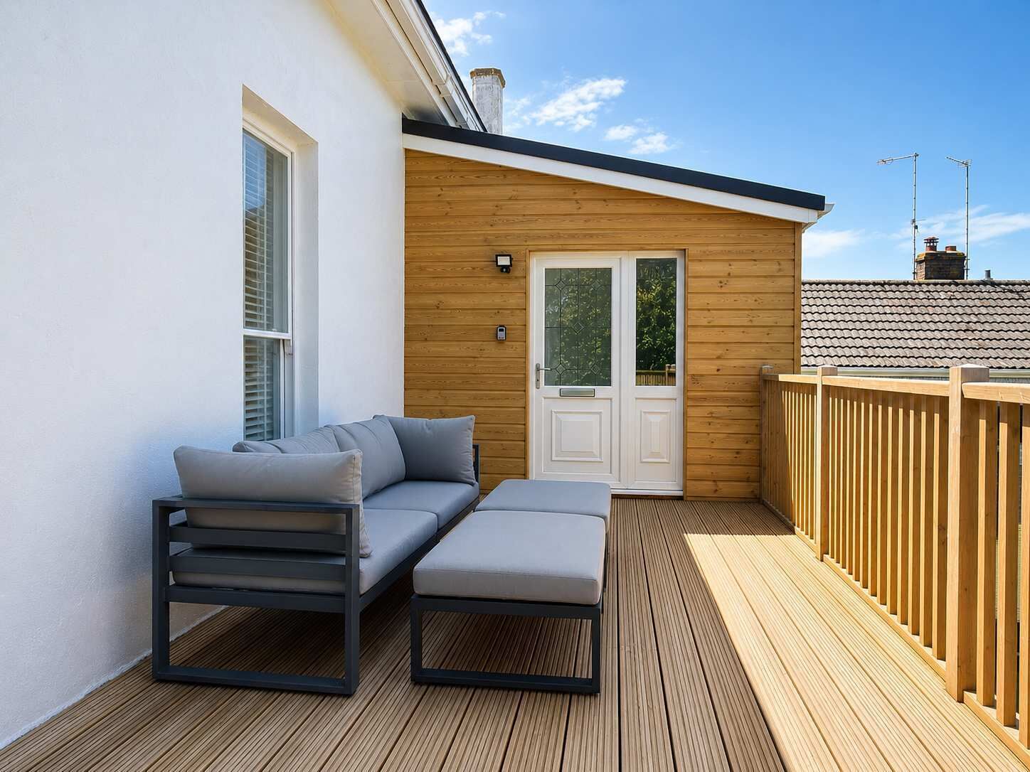 Une terrasse avec un canapé et un pouf, une porte en bois et un mur blanc.