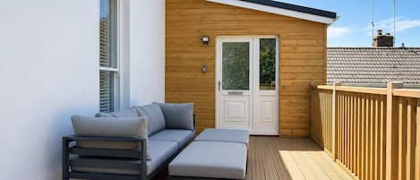 Une terrasse avec un canapé et un pouf, une porte en bois et un mur blanc.