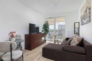 En moderne stue med sofa, fjernsyn på et træskab, spisebord med stole og en balkon med et planter.