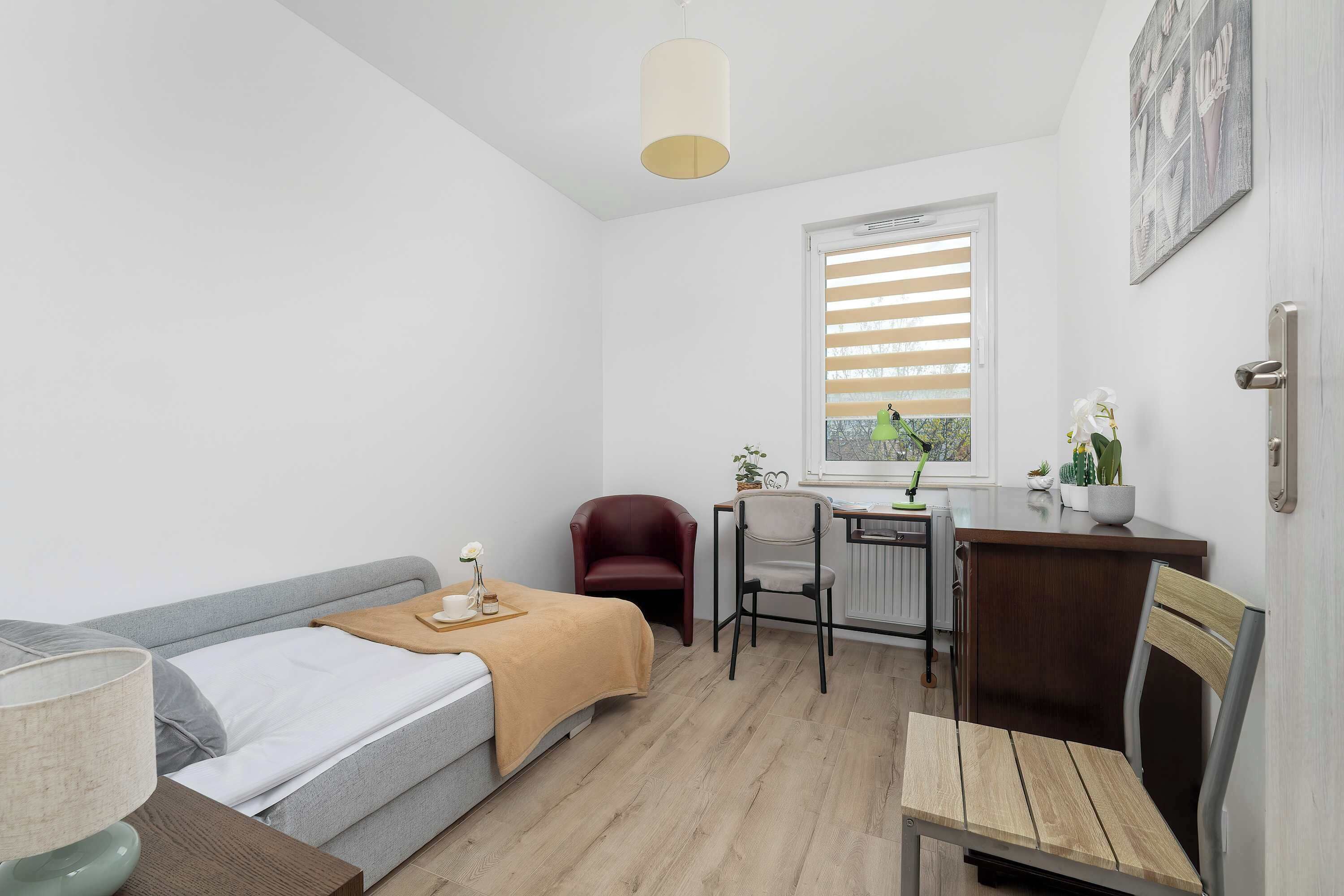 A small, well-lit room with a bed, a desk, a chair, and a window with blinds.