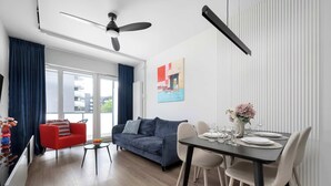 A modern living room with a sofa, dining table, and chairs. There is a ceiling fan, a window with curtains, and a painting on the wall. - Elegant Apartment with a View of the Oder River | Workspace | Parking | Balcony (Wrocław)