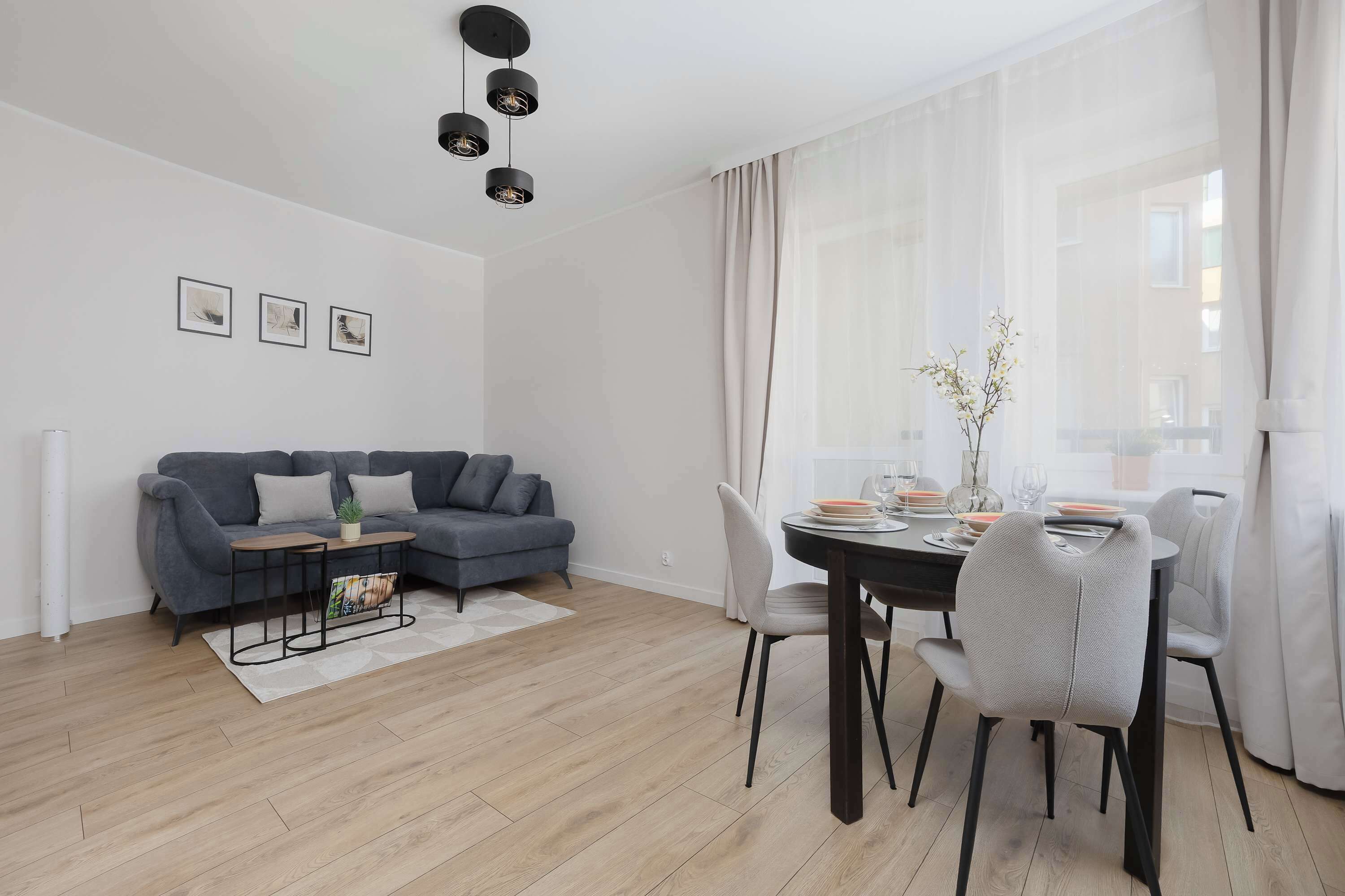 Una sala de estar moderna con un sofá gris, una mesa de comedor con sillas y una ventana con cortinas de gasa.