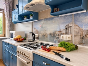 Cozinha com armários azuis, fogão a gás, micro-ondas e painel de azulejos com desenho de paisagem urbana.