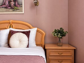 Una cama bien hecha con almohadas blancas y burdeos, cabecera de madera, mesita de noche con cajones, un jarrón con flores y una obra enmarcada en la pared.