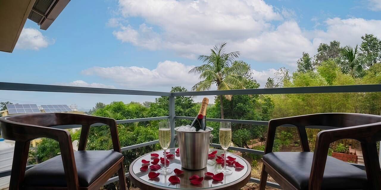 A balcony with a table set for two, a bottle of champagne, and red rose petals on a sunny day.