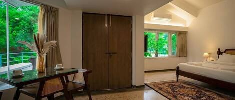 A hotel room with a bed, a desk, and a chair. There is a window with a view of greenery outside.