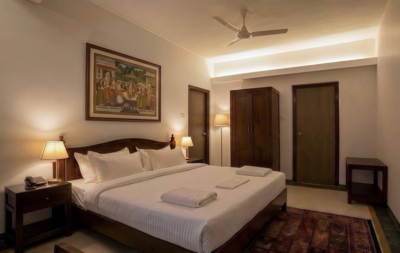 A hotel room with a bed, bedside lamps, a ceiling fan, a painting on the wall, and a door.